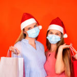female friends wearing surgical mask and Christmas hat holding shopping bags on a vivid red orange background